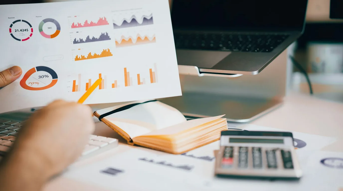 A hand lifting a printed chart sheet above an open notebook, a calculator and a laptop on the desk.