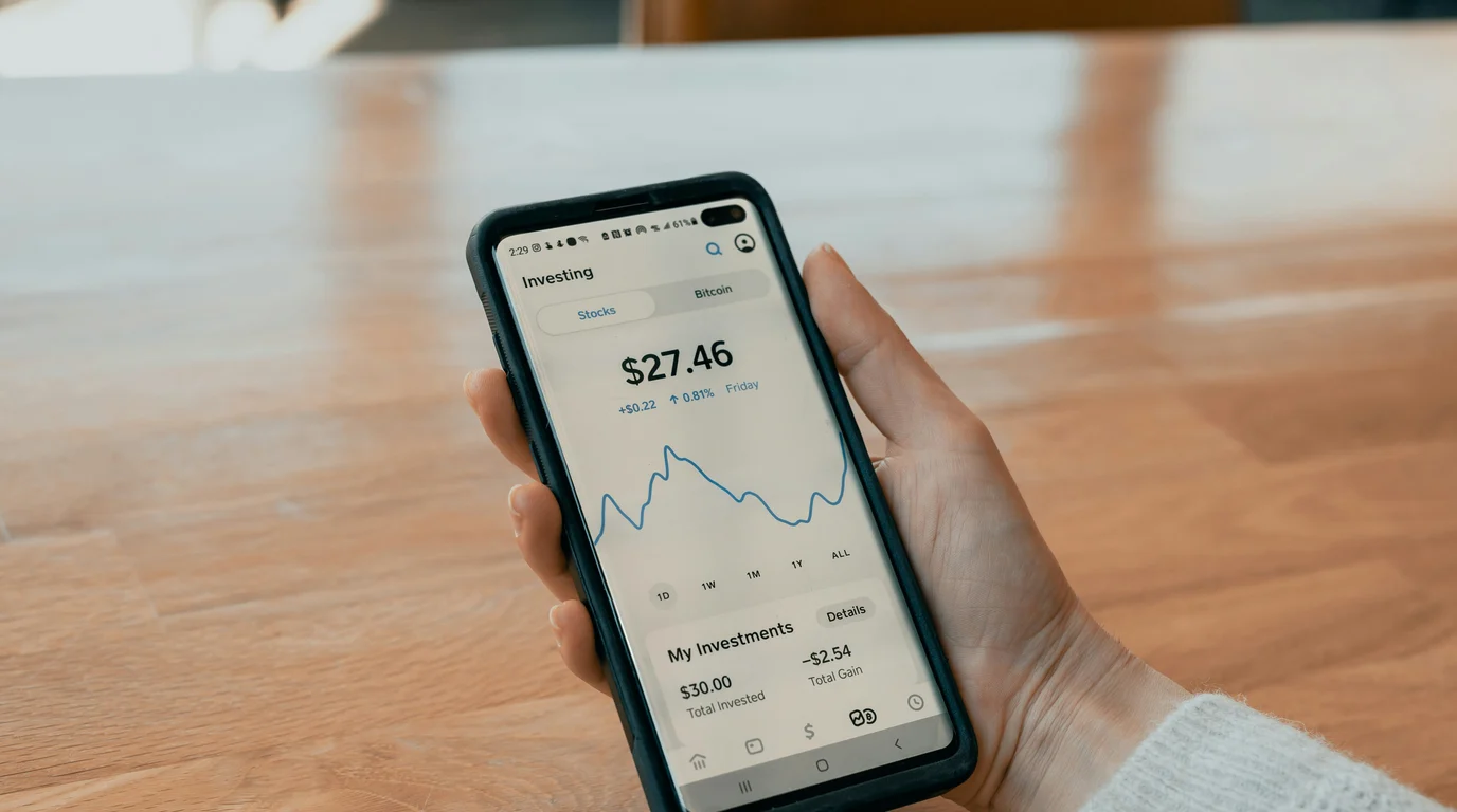A hand holding a phone showing an investing screen with a blue line chart and balance figures, above a pale wooden table.