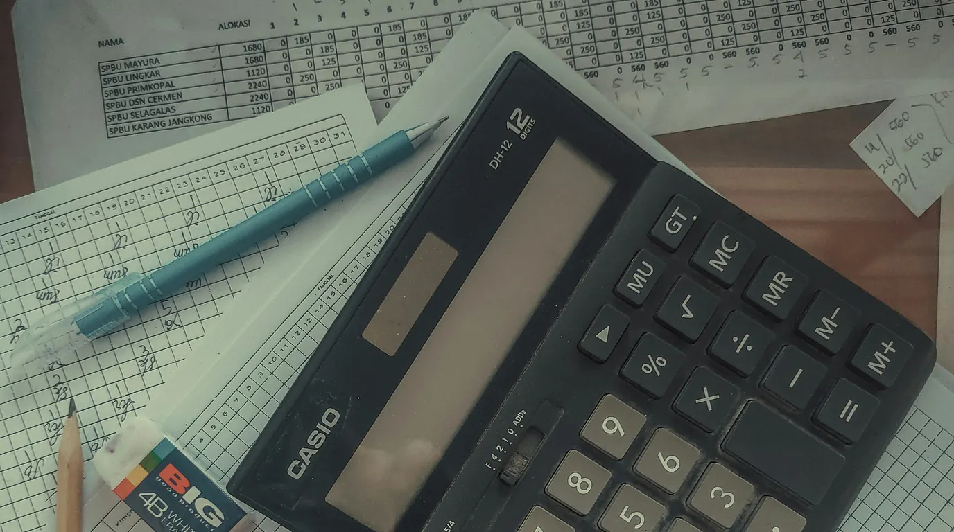 A black desk calculator lying across handwritten ledger sheets, with a blue mechanical pencil and an eraser nearby.