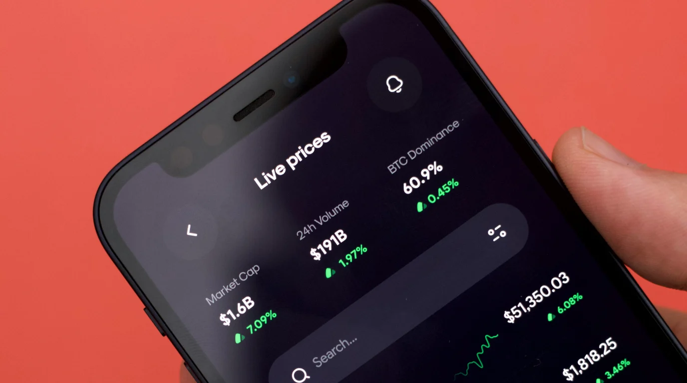 A phone held against a coral-red background, its dark screen listing live market prices and percentage changes in green.