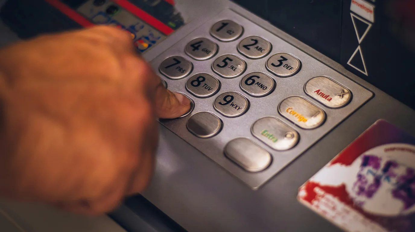 A thumb resting on the worn metal keypad of a payment terminal, orange and green function keys to the right.