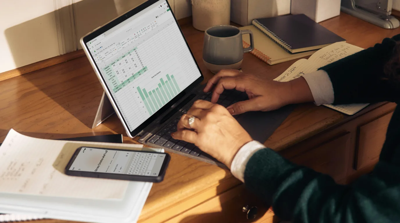 Hands working at a tablet showing green bar charts, a phone and notebook alongside.