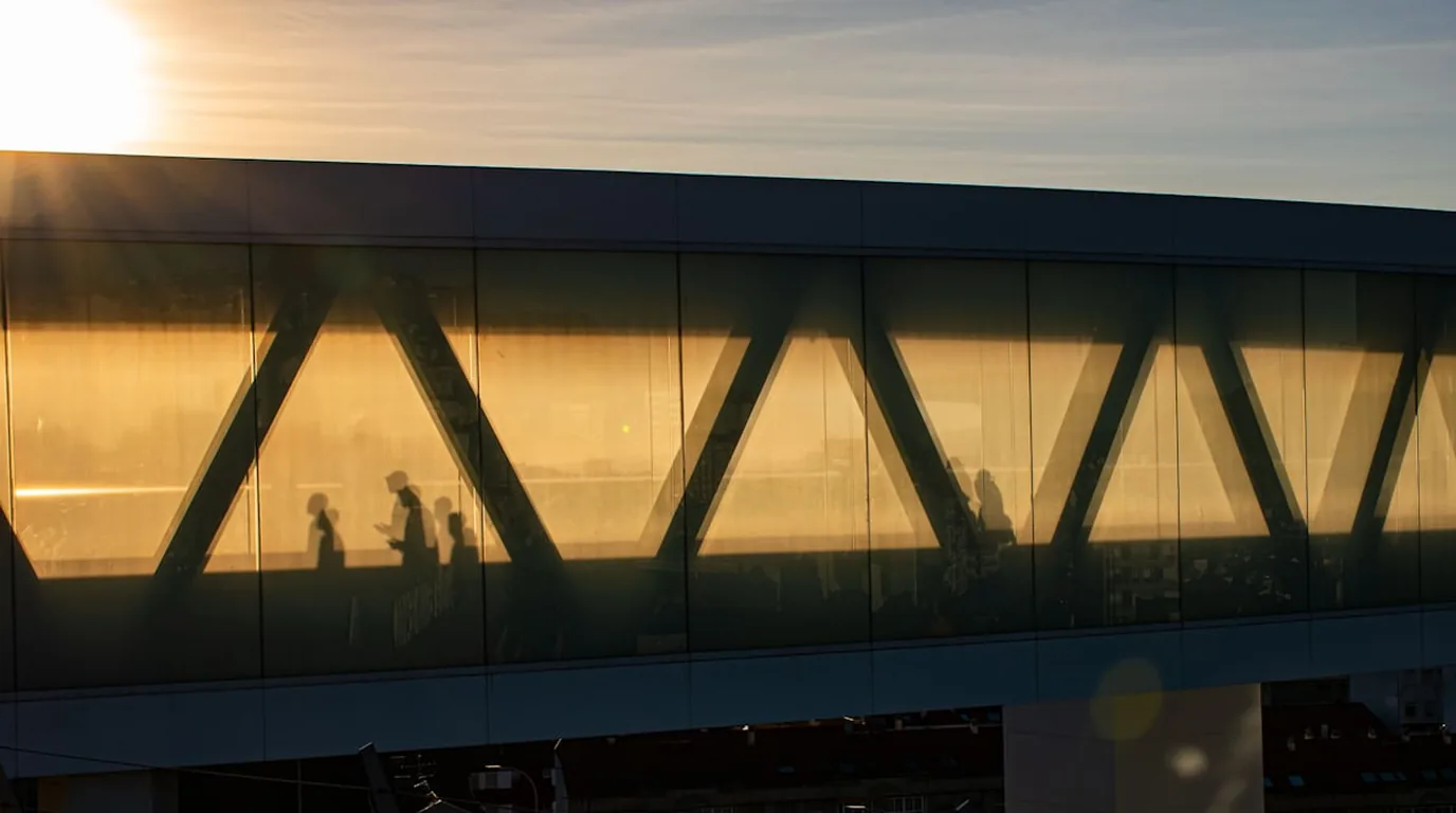 A glazed pedestrian bridge lit gold at dusk, figures crossing behind the glass.