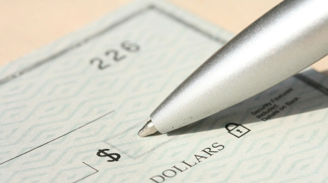 A silver ballpoint pen resting on a blank patterned cheque, close enough to read the printed dollar sign and amount line.