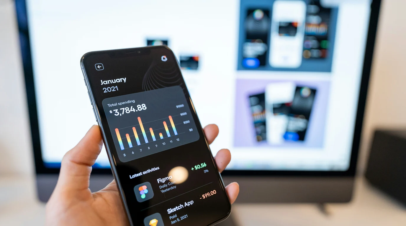 A hand holding a phone showing a dark spending dashboard with a colourful bar chart and a list of recent transactions.