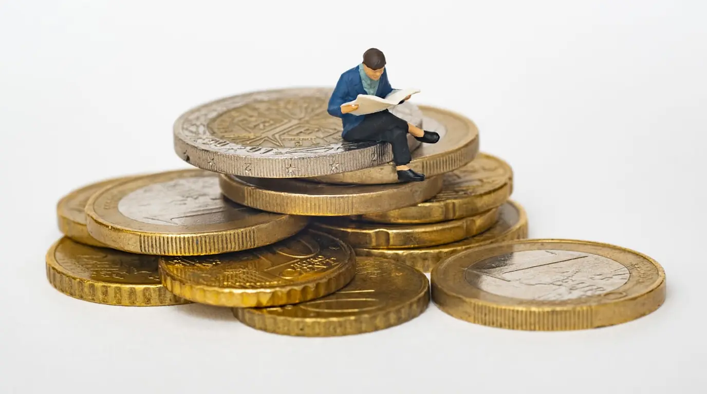 A miniature figure sitting reading on a stack of euro coins.