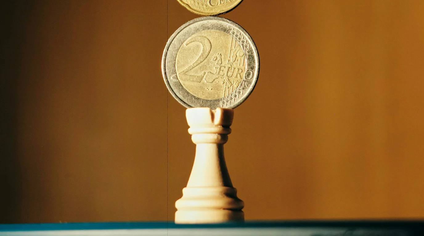A two-euro coin balanced upright on a white chess rook against a warm amber background, a second coin hovering above.