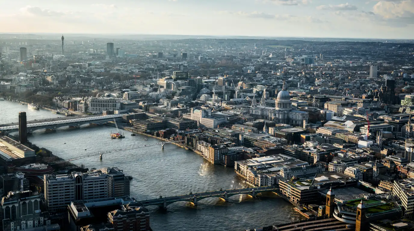 An aerial view over a wide river winding through a dense city, with bridges, a domed cathedral and rooftops stretching to the horizon.