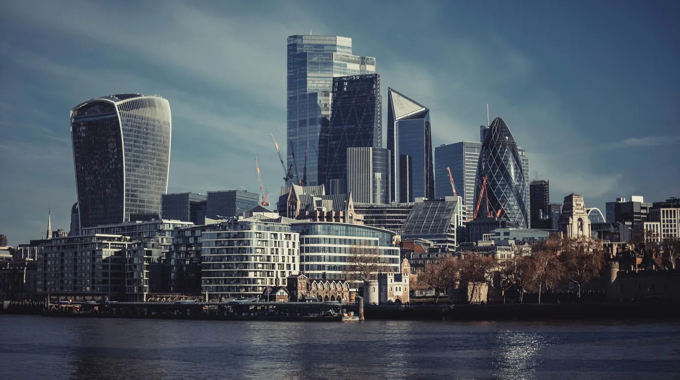 A skyline of glass towers in varied shapes rising above a riverside waterfront, seen across dark water under a blue sky.