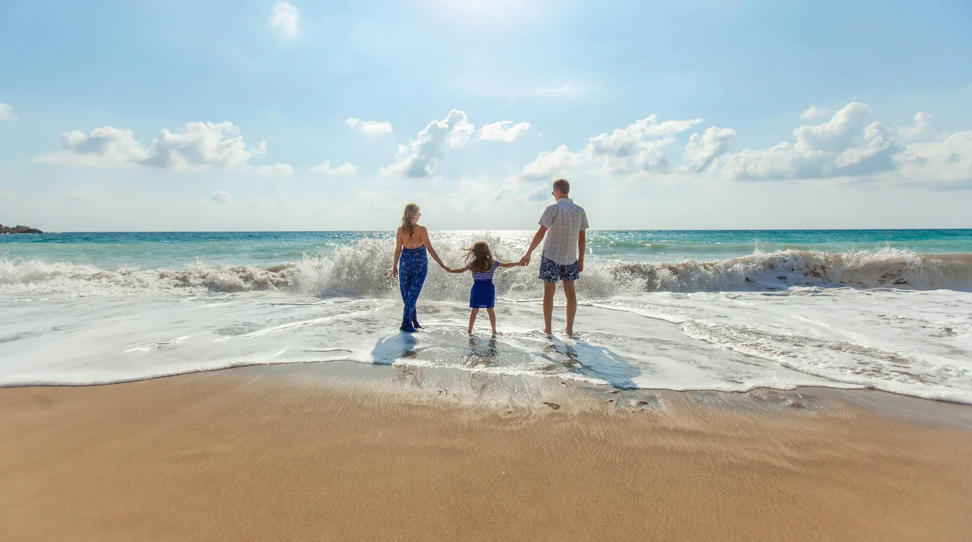 A family of three holding hands at the water's edge as a wave breaks around them.