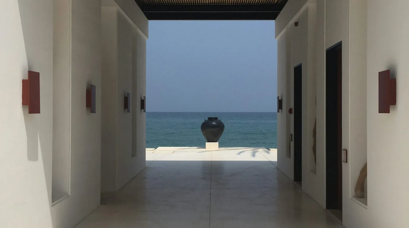 A white plastered corridor framing a dark ceramic jar on a plinth against the sea.