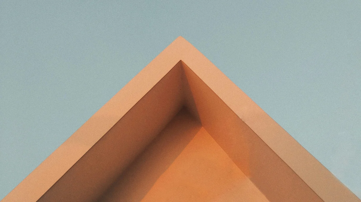 The sharp apex of a peach-toned concrete building cutting across a pale blue sky, its folded planes catching warm light.
