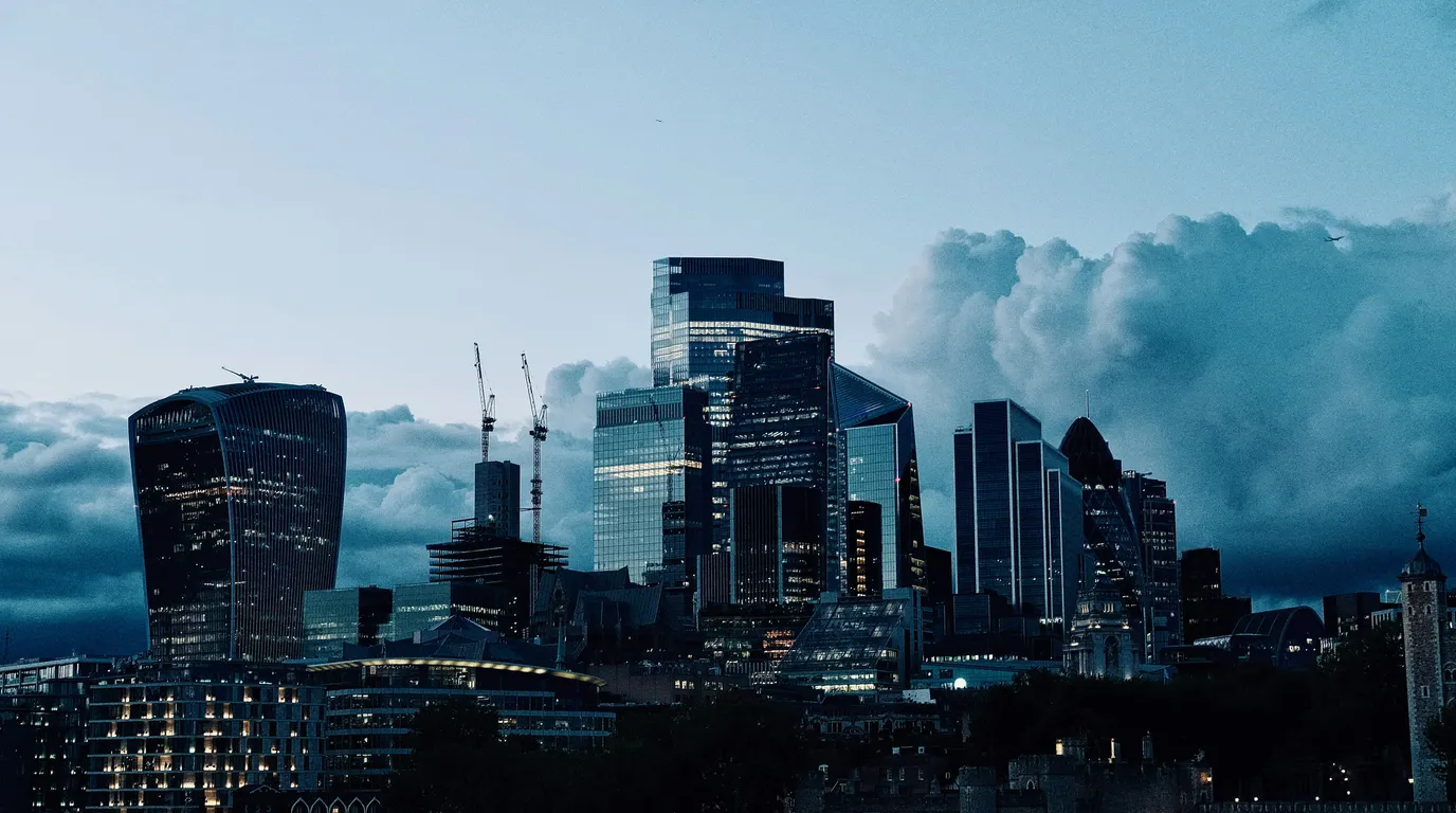 Lit office towers standing against a bank of storm cloud at dusk, the whole skyline washed in cool blue.