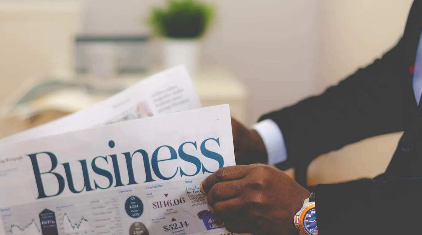 A person in a suit holding an open Business newspaper spread with market tables.