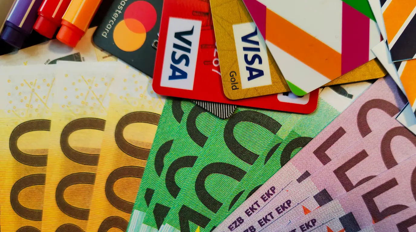 Payment cards in red, gold and black fanned above a spread of euro banknotes, with three marker pens at the top edge.
