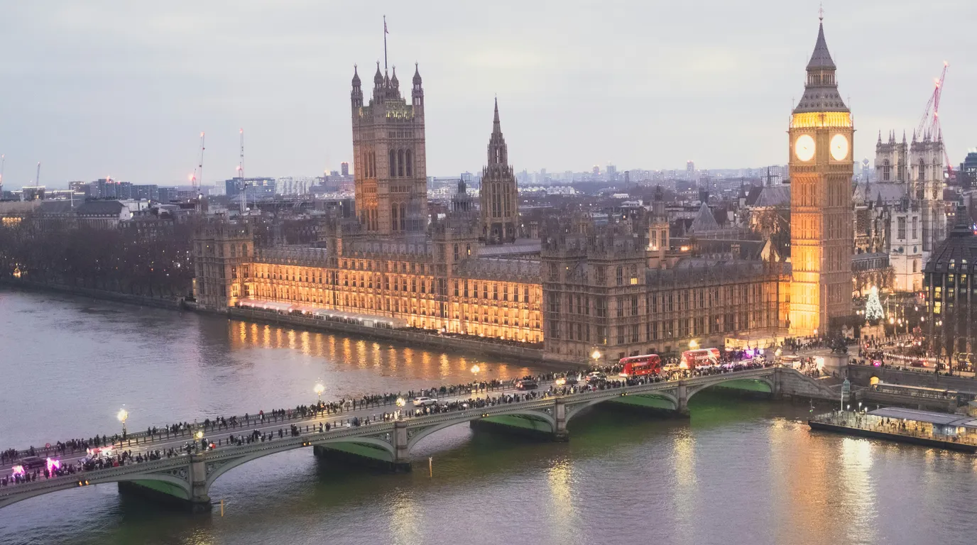 A floodlit riverside palace and clock tower glowing at dusk beyond a busy stone bridge crossing dark water.