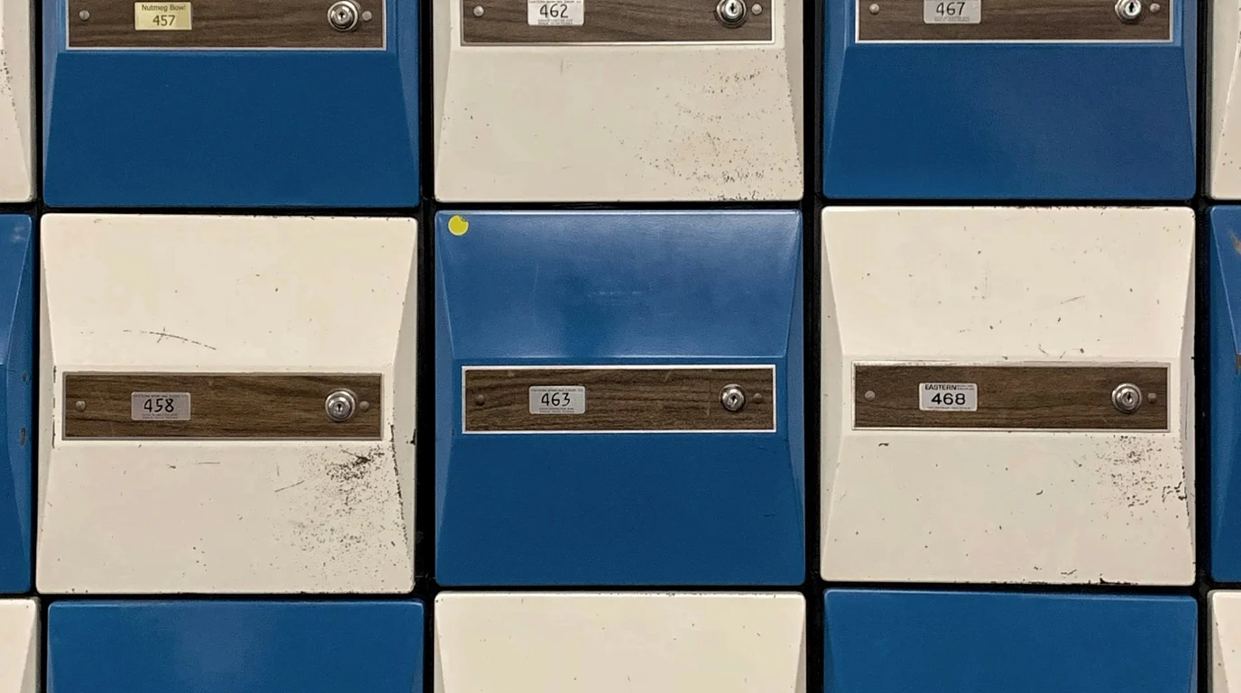 A grid of blue and cream metal locker doors, each with a small keyhole and a numbered plate set on a wooden strip.