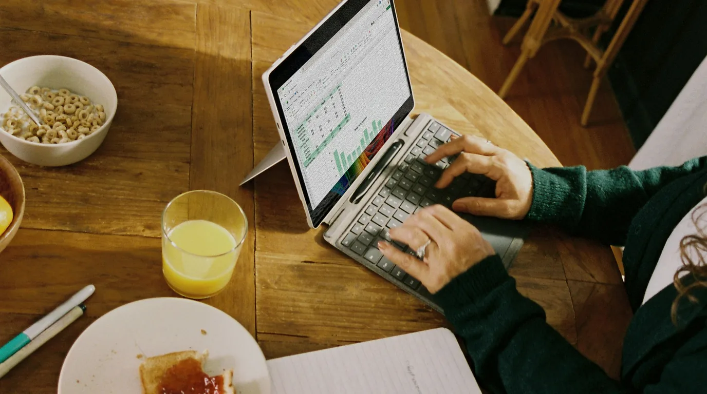 Hands typing on a laptop at a breakfast table, orange juice and cereal beside it.