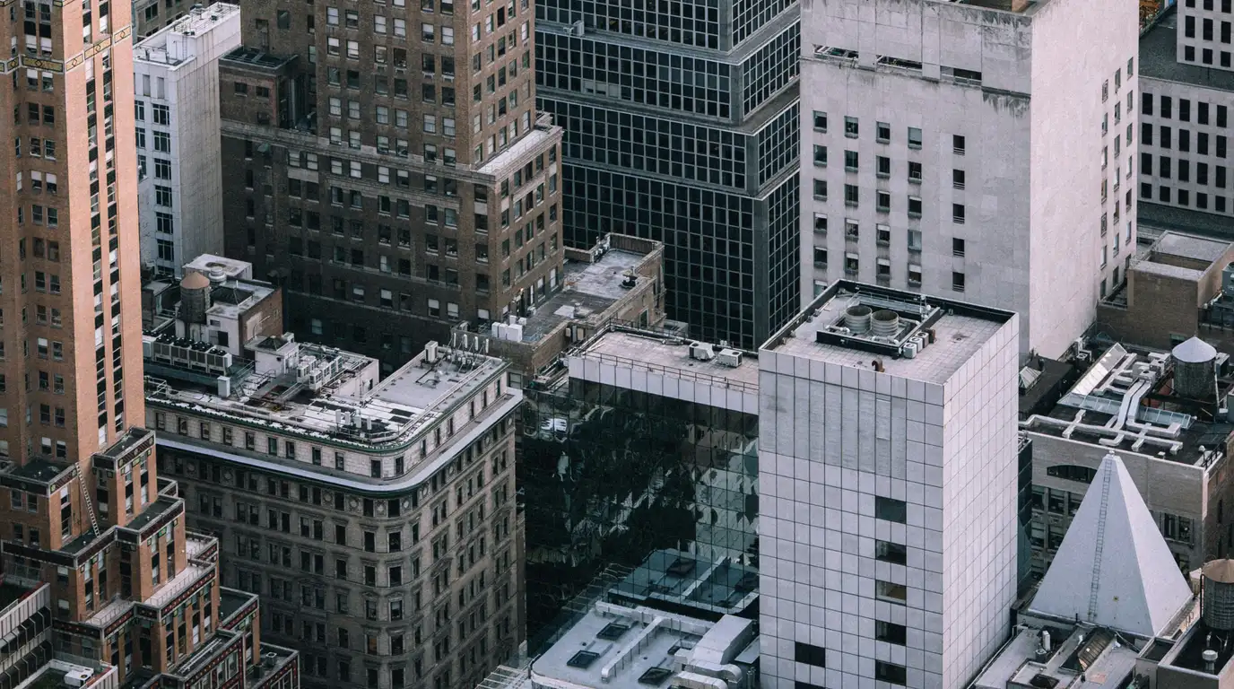 A high aerial view of packed city rooftops, where brick, glass and concrete blocks crowd together around rooftop machinery.