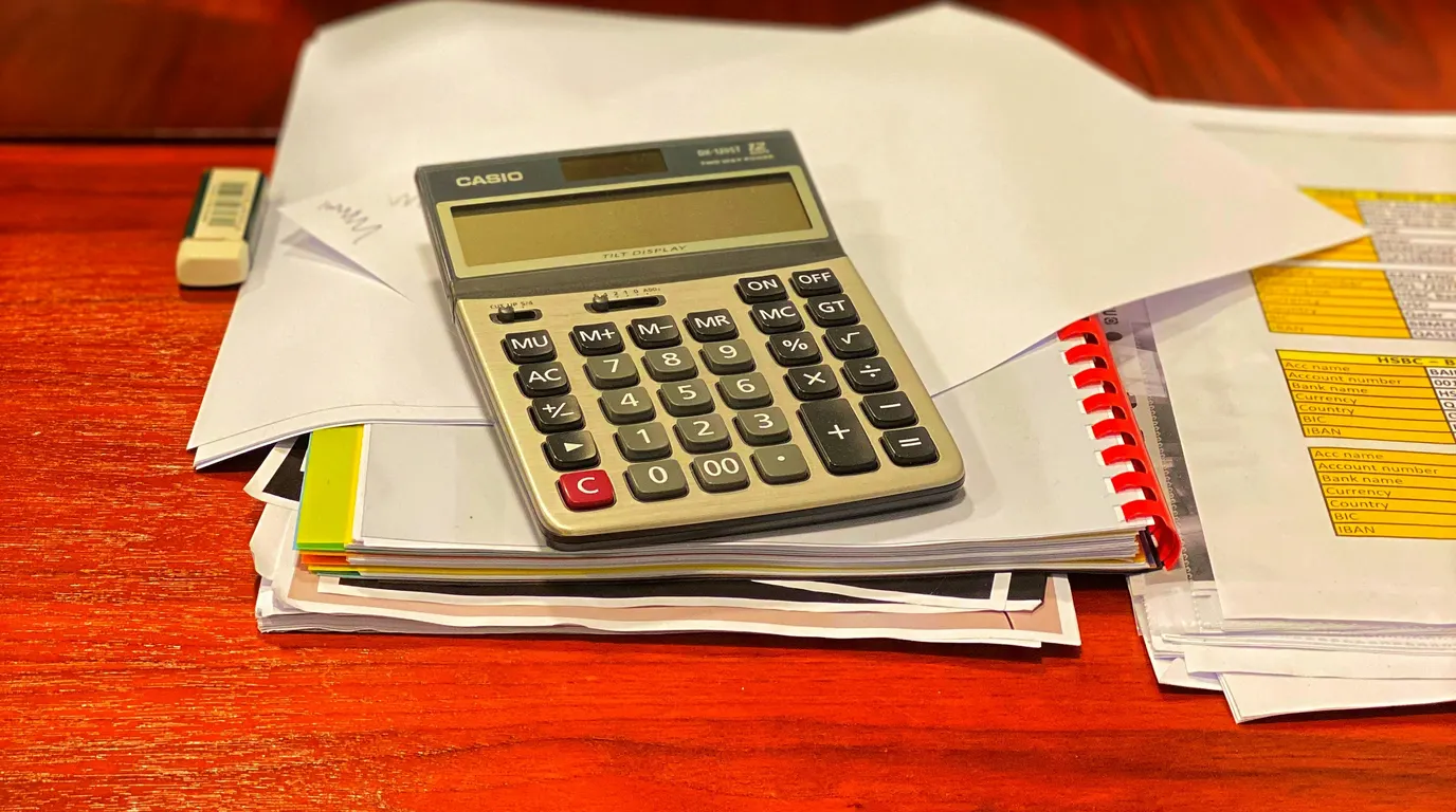 A large desk calculator sitting on a pile of loose papers and a spiral-bound document, on a glossy red-brown table.