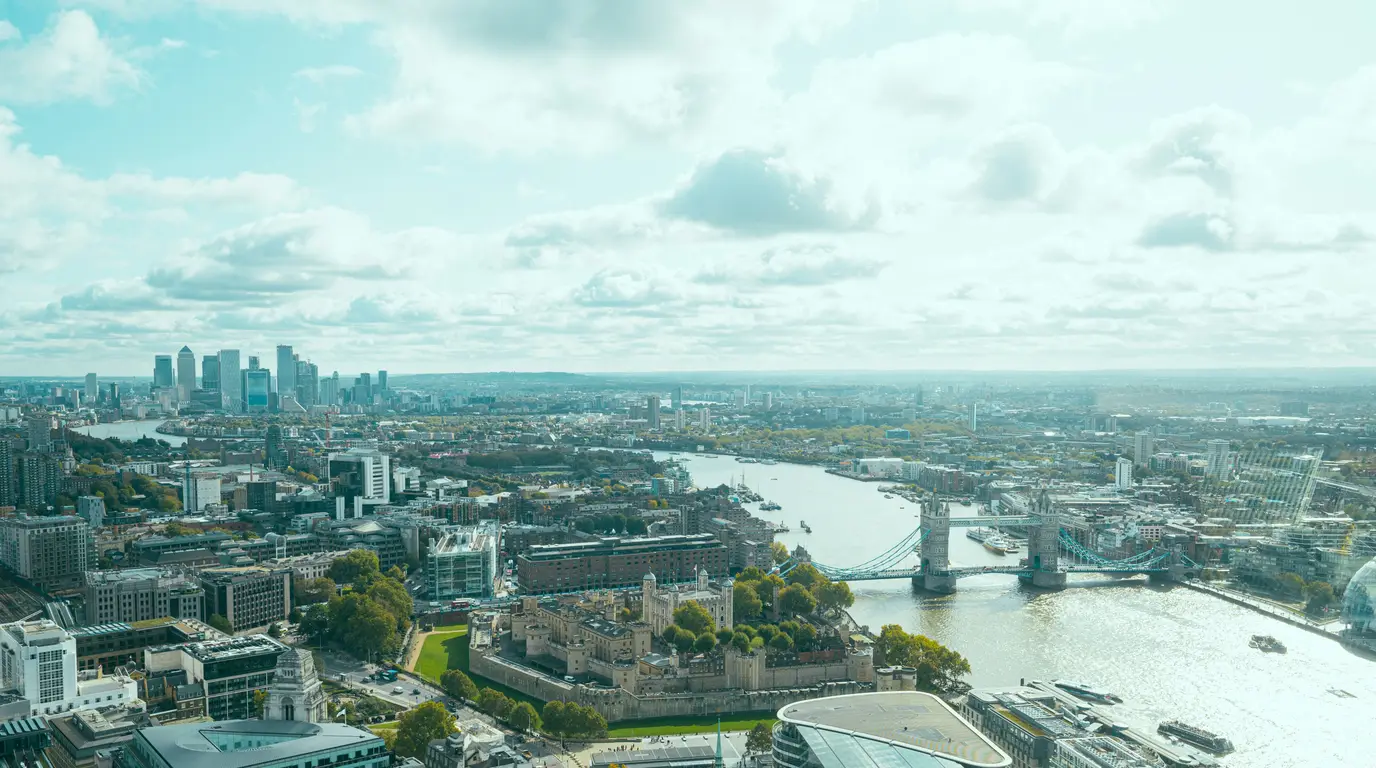 A hazy aerial panorama of a river winding through a city, with a twin-towered bridge and a walled fortress below.