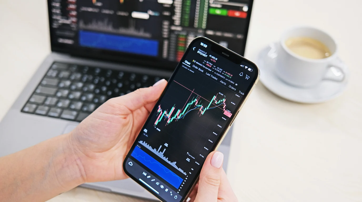 Two hands holding a phone showing a dark candlestick trading chart, with a laptop of market data and a coffee cup behind.