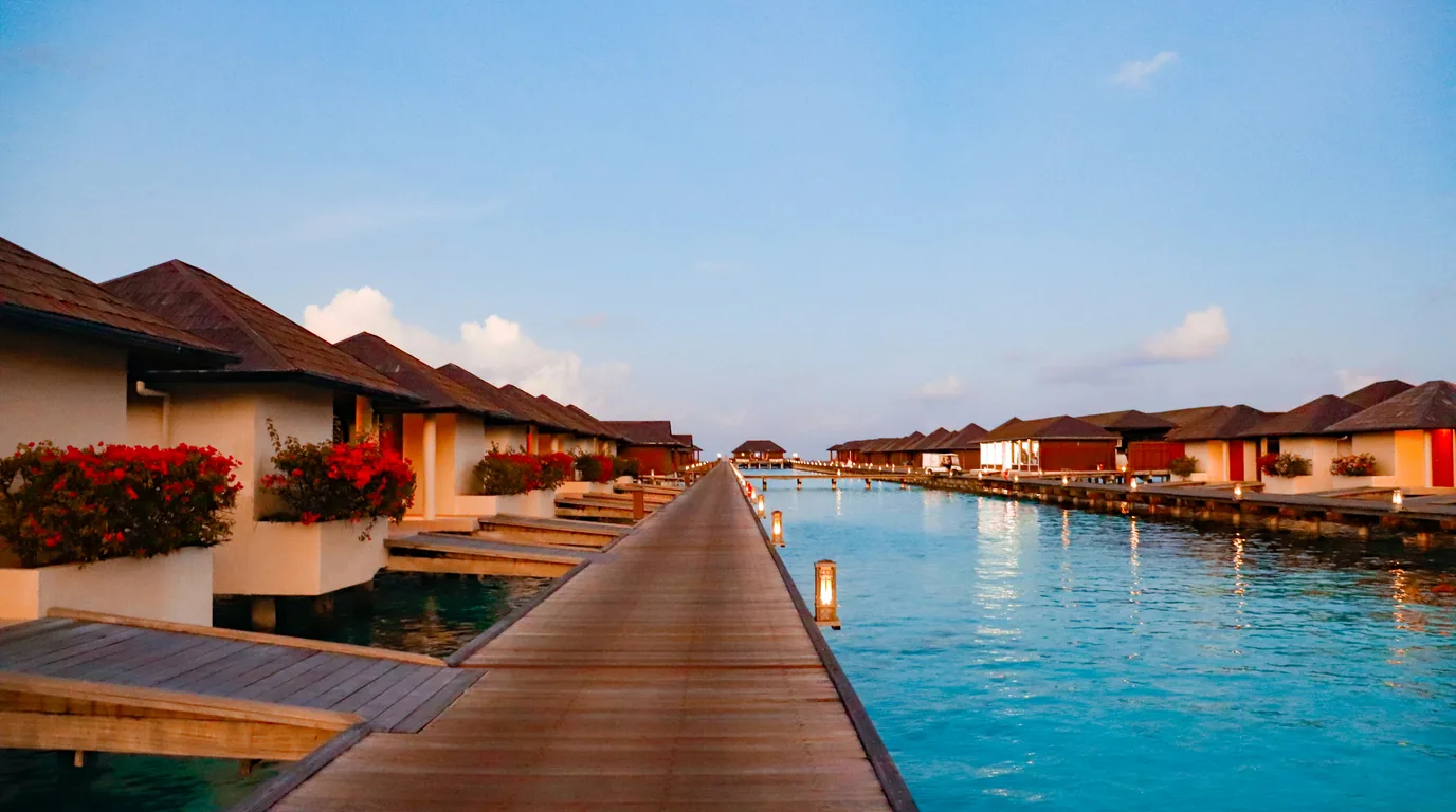 Thatched overwater villas at dusk, a lantern-lit timber walkway running out across still water.