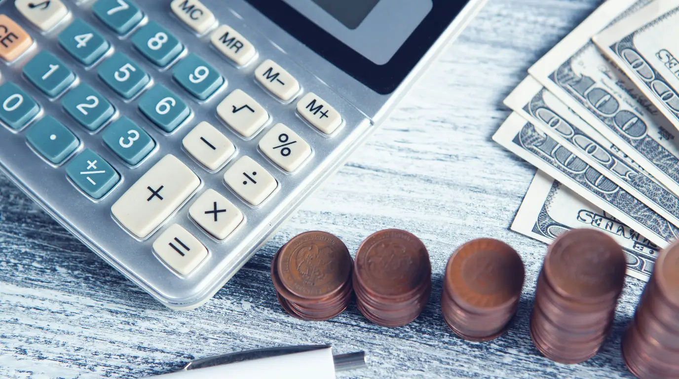 A calculator, a row of stacked copper coins and fanned banknotes on a pale weathered wooden surface.
