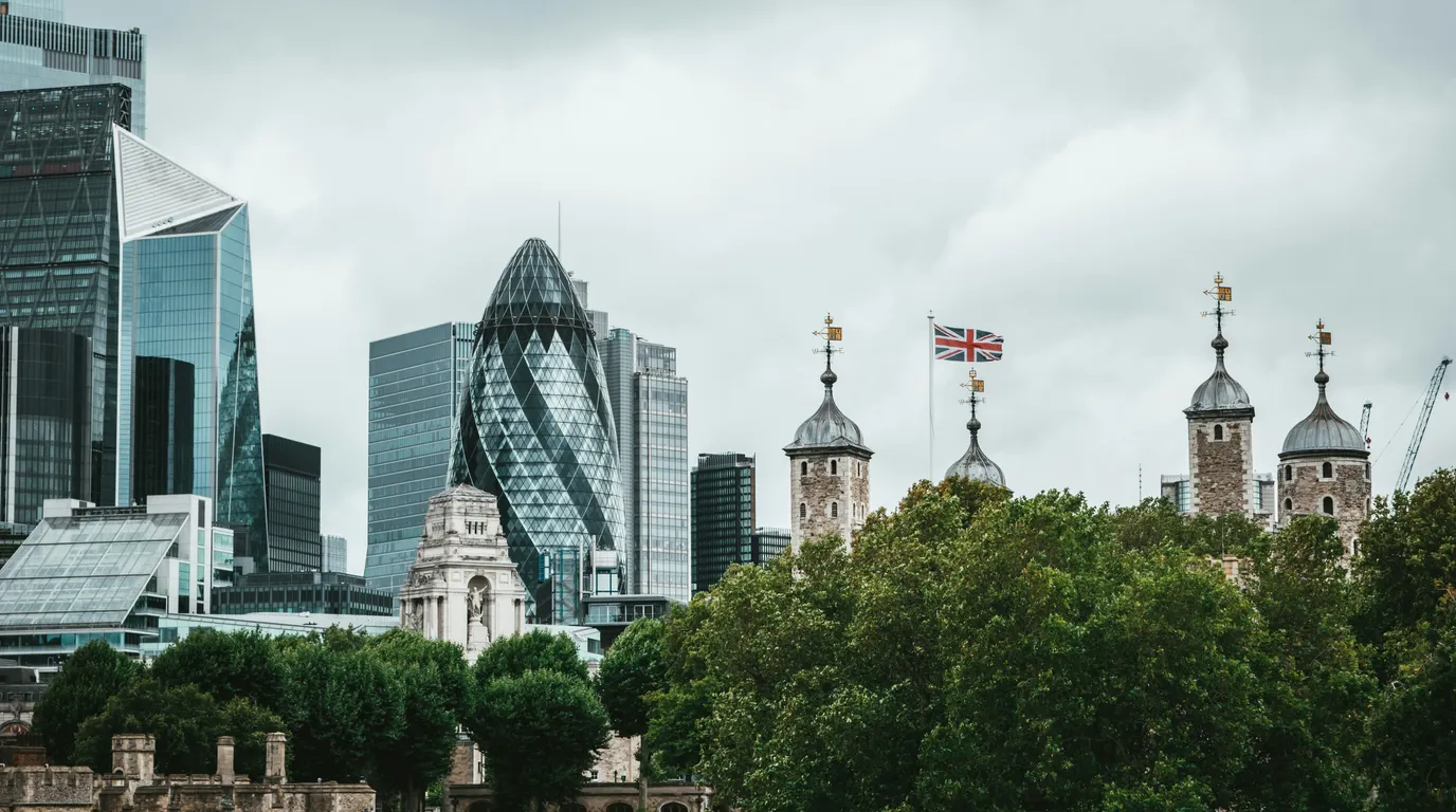 Glass office towers rising behind stone turrets and a fluttering flag, with dense green treetops below a flat overcast sky.