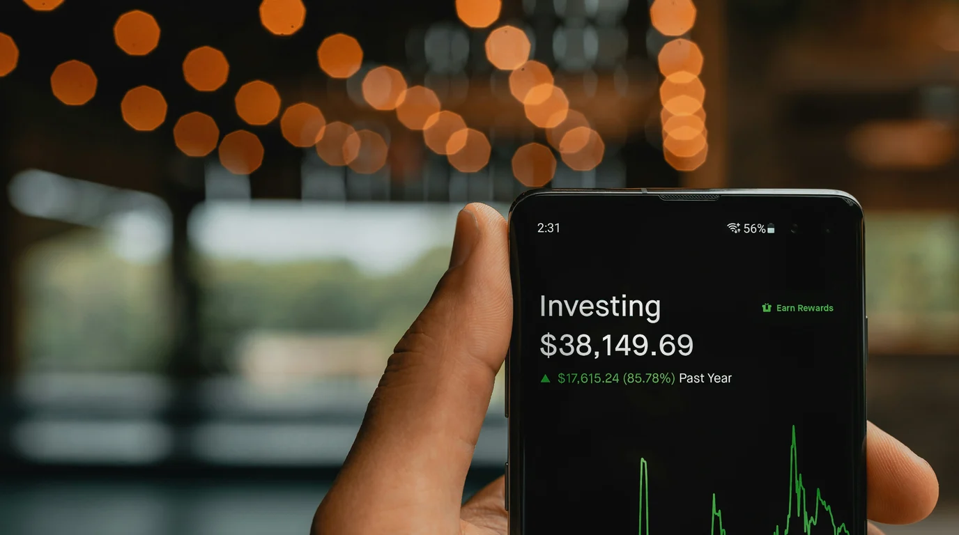 A hand holds a phone showing an investment balance and a green performance chart, with warm orange bokeh lights strung behind.