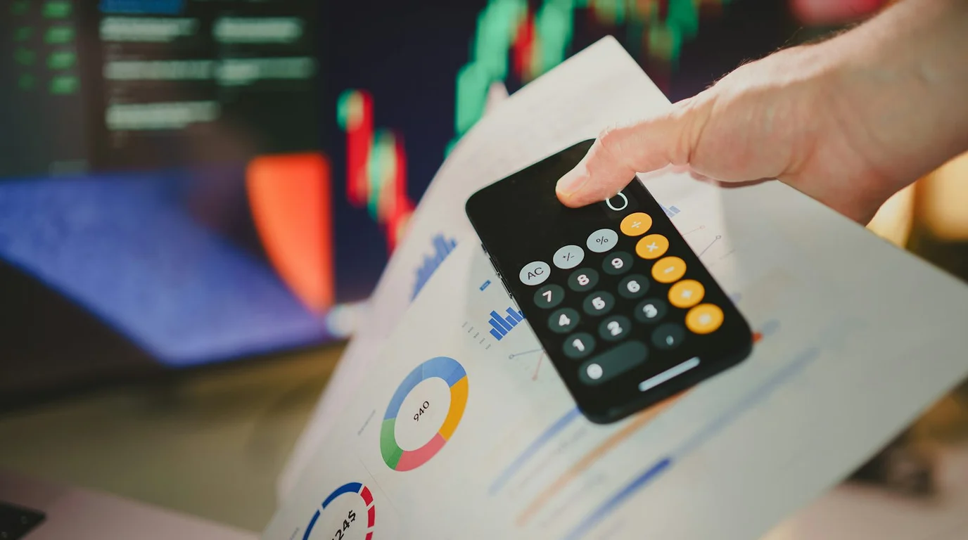 A finger reaches for a phone open on its calculator app, resting on printed pie and bar charts with blurred candlestick screens behind.