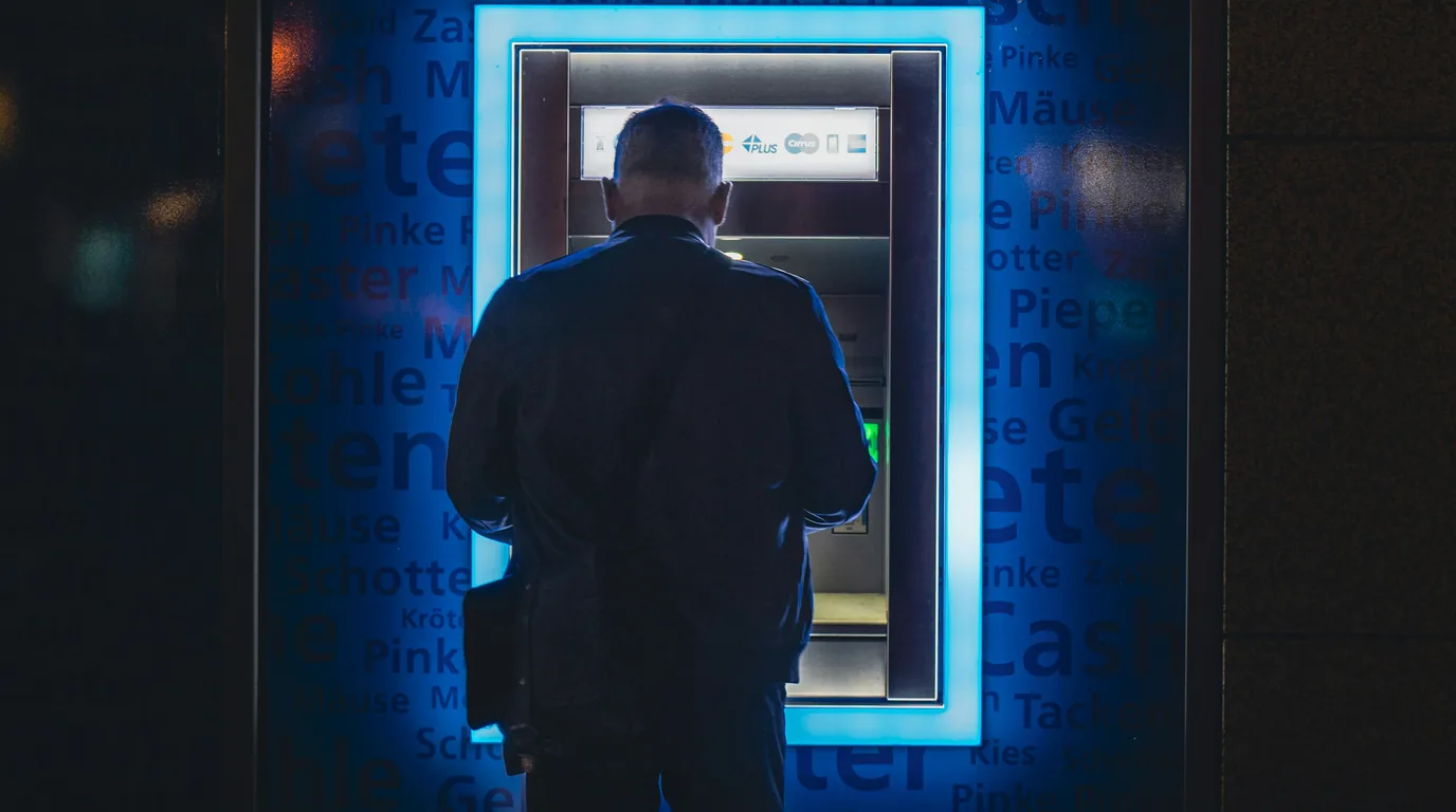A man at a cash machine in a blue-lit alcove, his back to the camera.